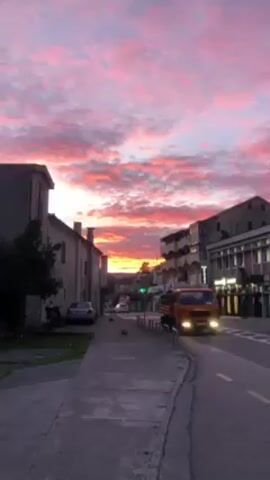 The video shows a street in a town at sunset. The sky is filled with bright pink and orange clouds. There are buildings on either side of the street, and a sidewalk runs along the left side of the street. A small orange truck drives on the right side of the road. Other cars are parked on the side of the street. The camera pans upwards to focus on the colorful sky.
