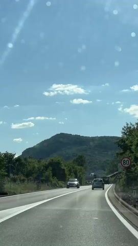 The video captures a forward-facing view from a moving vehicle on a multi-lane road in Barutana, Montenegro, on a bright, sunny day with a blue sky and scattered white clouds. The road has two lanes in the direction of travel, marked by white lines, and is flanked by lush green trees and foliage. In the background, a large, tree-covered mountain dominates the horizon.

Several cars are visible on the road, with a dark-colored car in the left lane and a white car in the right lane further ahead. Sunlight creates glare spots and reflections on the car's windshield.

Notable road signs include a circular speed limit sign on the right indicating "80" (likely km/h), and a triangular warning sign below it. Further down on the right, a white rectangular billboard is partially visible with the text "VOJTI POLOVNI DIJELOVI" (Used Parts). On the left side of the road, another billboard shows partial text like "DRIVA" and "BUS". Background music with a male vocalist is audible throughout the clip.