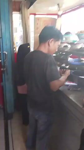 A man in a black t-shirt and blue jeans is standing behind a counter at what appears to be a food stall. He is holding a white cloth and looking down at something on the counter. Behind him, there are various bowls and containers of food. The stall is open-fronted with a red awning and a white roof. There is another person partially visible to the left of the man, wearing black clothing. The lighting is bright, suggesting it is daytime.