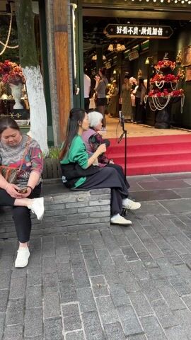 Two women are sitting on a stone bench in front of a store, filming themselves with a cell phone mounted on a tripod. The woman on the left has long, dark hair and is wearing a green sweater and black pants. The woman on the right has short, white hair and is wearing a floral top and black pants. Both women are looking at the phone screen. Another woman is sitting to the left of the main subject, using her own phone. The store behind them has a red carpet leading up to the entrance. There are other people visible in the background inside the store. The ground is paved with brick. A tree is to the left of the frame.