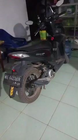 The video displays a black scooter parked indoors. The scooter is viewed from the right rear, with its license plate clearly visible. The background shows tiled flooring and a wall, along with various objects like a blue plastic chair, shelves with items, and a large blue water jug. The lighting is dim.