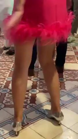 The video shows a woman from the waist down, facing away from the camera and dancing on a patterned tile floor. She is wearing a short, bright pink dress with a feathered hem and silver, pointed-toe heels. Other people's feet are visible around her, suggesting she is dancing in a group.