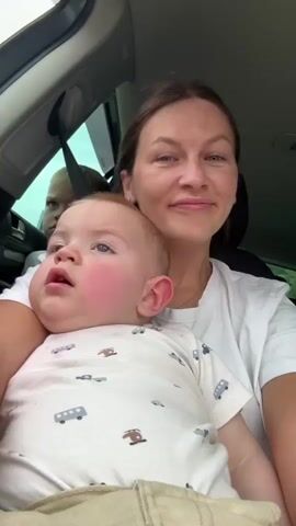A woman in a white t-shirt is in the front passenger seat of a car, holding a baby in her arms. The baby is wearing a white onesie with a pattern of cars. The woman kisses the baby on the forehead. In the background, visible through the side window, a child in a booster seat is looking out the window. The car appears to be moving, as indicated by the slight motion blur.