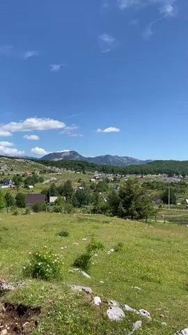 A drone flies across a clear blue sky over a scenic valley dotted with houses. The valley is filled with green grassy fields and trees. Mountains can be seen in the background. The drone is visible as a small black dot in the upper right quadrant of the frame. The video pans slightly to the right, showing more of the valley and houses. The scene is peaceful and idyllic.