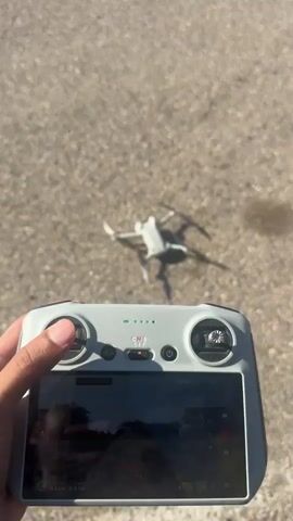 The video shows a drone being operated outdoors. In the first few seconds, a person's hands are visible holding a drone controller, with the drone itself resting on a textured ground surface in the background. The drone then takes off and ascends into the air. The subsequent frames capture the drone flying against a clear blue sky. In the background, a building with a red roof and a yellow and purple van parked in front of it are visible. Several people are seen on the roof of the building.