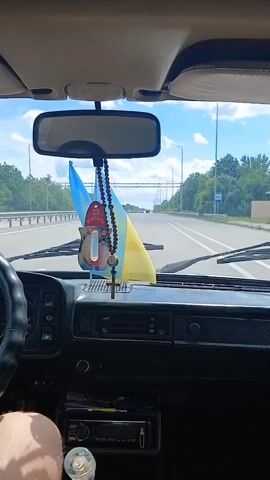 The video shows the interior of a car, looking out through the windshield. The car is in motion on a highway. On the dashboard, there is a rearview mirror hanging from the ceiling. Draped from the rearview mirror are a rosary with a cross and a small Ukrainian flag. The Ukrainian flag is blue and yellow. The road ahead is visible, with trees lining both sides. The sky is cloudy. The dashboard of the car is visible, with a steering wheel on the left and various controls and a radio. A person's leg is visible in the lower left corner of the frame, suggesting they are the driver or passenger. The video is taken from inside the car as it travels down the highway.