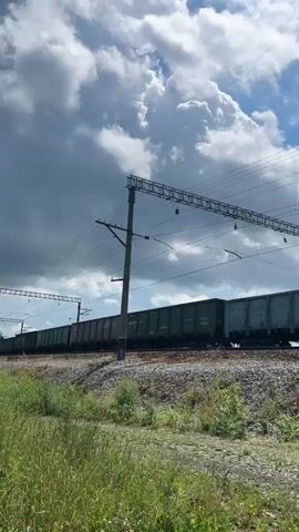 A freight train moves from left to right across the screen. The train consists of multiple open-top hopper cars, all dark gray. The cars are numbered and appear to be carrying some material, possibly ballast or aggregate, as their contents are indistinct and appear to be at a level below the rim of the cars. The train is moving on a track bordered by gravel and green vegetation, including tall grass and some smaller plants. Overhead, electrical lines are visible, supported by poles and a network of wires, indicating an electrified railway. The sky is blue with scattered white clouds, some of which are large and puffy. The overall impression is a daytime scene on a sunny day, with the train in motion along a rural or semi-rural route.