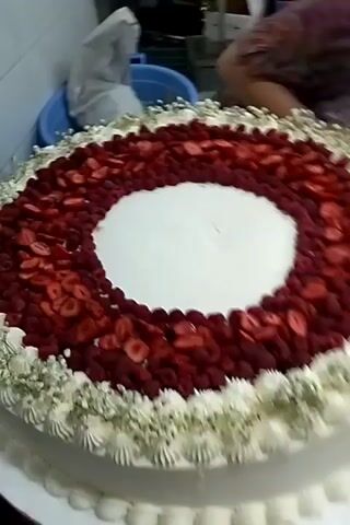 A large, round, white cake is displayed on a white platter. The cake is decorated with a wreath of red berries, possibly strawberries and raspberries, around a circular white center. A border of white frosting with small white flowers encircles the berry wreath. The side of the cake is decorated with piped white frosting scallops and a red ribbon tied in a bow on the right side. The background shows a kitchen environment with a tiled wall, shelves, and some boxes. A person in a purple garment is partially visible in the upper right corner, appearing to be arranging something. The camera is focused on the cake, with a slight rotation to the right.