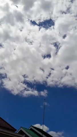 A video shows a close-up view of a bright blue sky with scattered white clouds. The clouds are puffy and appear to be moving slightly. The bottom of the frame shows the top of a building with a dark, curved edge. A thin, dark vertical pole with a horizontal element at the top is also visible extending upwards from the building into the sky.
