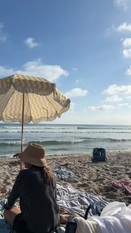 The video shows a beach scene on a sunny day with some clouds. In the foreground, there are several beach chairs and towels spread out on the sand. A yellow and white striped umbrella is set up. Further back, the ocean waves are rolling in. There are a few people scattered on the beach, some sitting in chairs and others standing. Two palm trees are visible on the right side of the frame. The sun is bright, casting a glare in the sky.