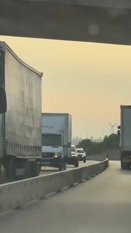 The video shows a highway scene at dusk. A white box truck with the logo ANTON on the side is in the left lane. In the right lane, there are two dark-colored cars following each other. In the distance, several wind turbines are visible against a hazy, orange-tinted sky. The road has concrete barriers on the right side, and there are trees and hills in the background. The video progresses to focus on a white car driving on a curved section of the highway, with wind turbines continuing to be visible in the distance.