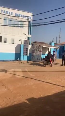 The video shows a scene in front of a building labeled "D.F.F TABLE WATER LTD." The building is white with a blue stripe. A motorcycle rider in a black jacket and pink pants is near the entrance where three people are standing, two in suits. A small structure with a sign sits between the motorcycle and the building. The road is dirt or gravel. Utility wires are visible overhead.