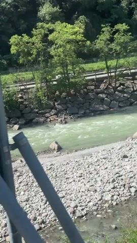 The video shows a wide, shallow river with greenish, turbulent water flowing over a rocky bed. The riverbanks are lined with a mixture of large rocks and smaller pebbles. On the left side, a steep, tree-covered hillside rises, with a road running along its base. Trees with lush green foliage are visible. On the right side, a similar pebbly shore leads to a more distant, gently sloping hillside with some buildings and vegetation. A concrete bridge with visible support pillars spans the river in the distance, and there appear to be some vehicles on or near the bridge. The overall environment is natural and appears to be a scenic outdoor location.
