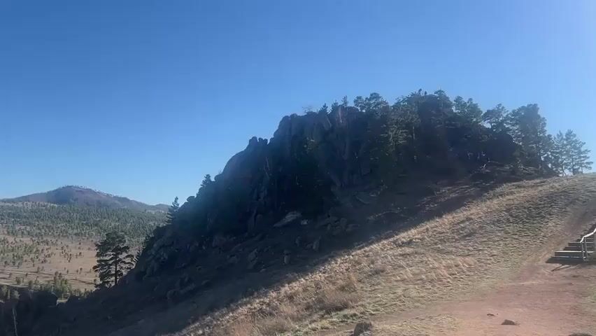 The video shows a rocky hill under a clear blue sky.  The hill has trees growing on top of and along its sides.  The ground in front of the hill is covered with dry grass.  In the distance, there is a mountain range covered with trees and some snow on the peaks.  A set of stairs is visible on the right side of the frame.