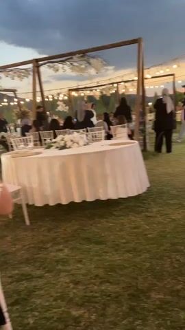 The video captures an outdoor event, possibly a wedding reception, at dusk. In the foreground, a round table draped in a white tablecloth is set on a grassy area. The table has a floral centerpiece. White chairs surround the table. In the midground, a wooden archway decorated with white foliage is visible. Several people are gathered, some standing and some seated. A woman in a black outfit and a light gray hijab is facing away from the camera, standing near the archway. Other guests, some wearing formal attire, are scattered throughout the scene. String lights are hung overhead, casting a warm glow. In the background, trees and hills are visible against a dusky sky. The overall atmosphere appears festive and relaxed.