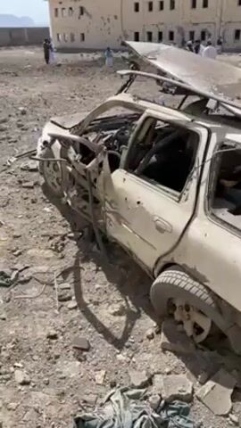 The video shows a severely damaged white station wagon in a desolate, rocky landscape. The car's roof is partially detached and bent, and there are numerous bullet holes and dents throughout the body. The windows are shattered, and the interior is visible, though it's difficult to discern details. In the background, there's a building with several windows, some of which appear to be broken. A group of people, mostly wearing traditional Afghan clothing, are standing near the building. The environment suggests a conflict zone or a location that has experienced significant violence. The overall tone of the scene is somber and indicative of destruction and potential loss of life.