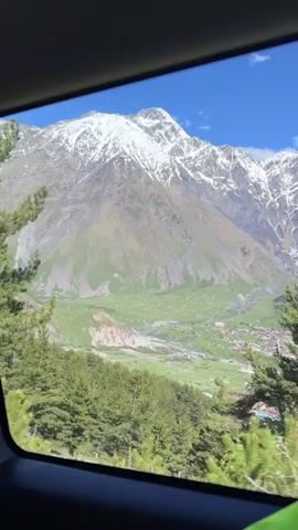 The video shows a view from inside a moving car, looking out the window. The landscape outside consists of pine trees and distant mountains, partially covered in snow. The sky is blue.