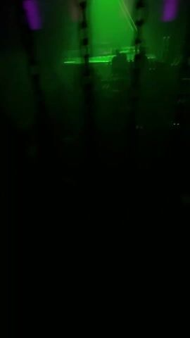 The video shows a crowd of people in a dimly lit nightclub, viewed through several chains hanging from the ceiling. The scene is illuminated with green and purple lights, and a DJ can be seen on a stage in the background. The silhouettes of people are visible dancing and moving to the music. The atmosphere is energetic and lively.