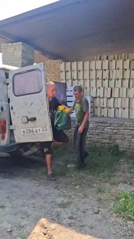 Two young men are by the open rear doors of a white van. The man in the foreground, wearing a black t-shirt with a graphic print, black shorts, and black sandals, holds a green bag with a yellow inner pouch. He is facing the camera and looking to his left. The man in the background, wearing a green t-shirt and dark pants, is partially inside the van, appearing to reach for something. The van is parked on a dirt surface. Behind the men, a stone-clad building with a corrugated roof is visible. The building has stacks of light-colored, rectangular objects, possibly boxes or crates, against its wall. The environment appears to be outdoors in daylight, with vegetation and dirt visible.