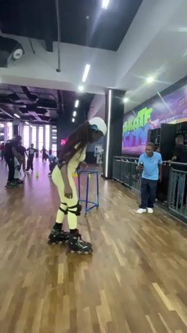 In an indoor skating rink brightly lit by overhead fluorescent lights, a dark-haired woman in light yellow athletic wear and a white helmet practices roller skating. She is holding onto a blue metal walker for support. To her right, a man in a blue shirt stands behind a low black fence, holding a phone, possibly recording her. Behind him, another man in a black shirt stands. In the background, other people skate around orange cones. A large mural with the words "Skate City" adorns the wall to the right. The floor is wooden, and in the distance, a neon sign reading "Roll with Us" is visible.