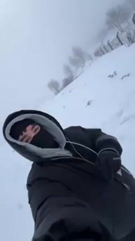 A person in a black ski mask and black gloves stands on a snowy hill. A fence is visible on the hilltop. The person holds up a hand for a moment. The camera then pans over a group of people walking on a snow-covered field beside the fence. A cloudy, foggy, overcast sky is overhead. The ground is covered in deep snow. Footprints are visible in the snow.