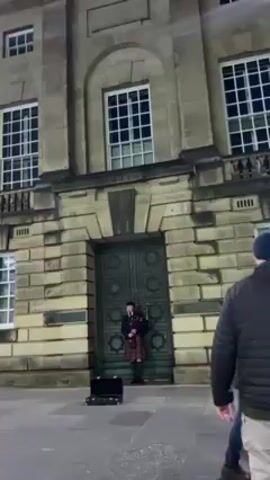 A medium, low-angle shot depicts a man playing bagpipes in front of a large stone building with multiple windows. The man is dressed in traditional Scottish attire, including a kilt, and stands in front of a large, arched doorway. A black instrument case sits on the ground near him. A man in a black jacket and khaki pants walks by on the right side of the frame. The building is made of large, light-colored stone blocks, and the lighting suggests it is evening.