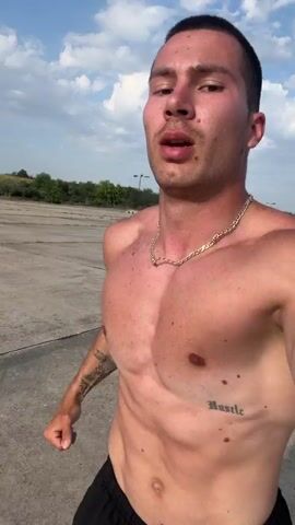 A shirtless, muscular man with short dark hair, a gold chain, and visible tattoos (including "Hustle" on his left rib cage and a design on his left bicep) is shown from the chest up, moving forward in an outdoor, paved area. He appears to be jogging or power-walking, looking forward and slightly to the left, with his mouth slightly open as if breathing heavily. The background consists of a large, empty concrete or asphalt surface under a partly cloudy, bright blue sky, with some green foliage and a distant light pole visible.