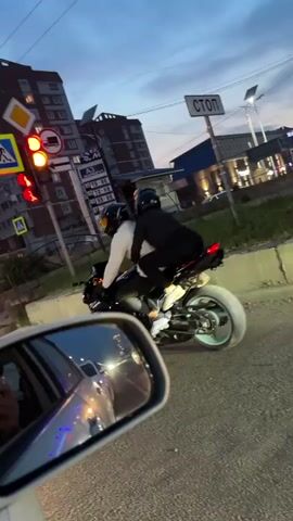 A motorcycle carrying two people, both wearing helmets, turns at an intersection. A car's side mirror, reflecting city lights and buildings, is in the foreground. Traffic lights are visible, with one showing green. The sky is dark, suggesting it is evening or dusk. Tall apartment buildings are in the background.