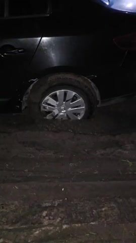 A black car is stuck in mud at night. The camera pans from the rear of the car, showing one wheel deeply embedded in mud, to the front of the car, where the headlights illuminate the muddy terrain. The car's doors and side panels are visible, as is the interior through the windows. The ground is covered in dark, wet mud, with tire tracks visible. In the final shot, a person is standing behind the car.