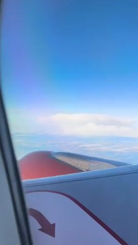 The video shows an aerial view from an airplane window. The camera is positioned to capture the airplane's wing, which is prominently featured in the foreground. The wing is predominantly white with a red stripe along the edge and a red arrow pointing downwards on the white surface. Part of the engine nacelle, colored red and silver, is visible. Beyond the wing, a vast expanse of blue sky stretches out, with a band of white clouds forming a line across the horizon. Below the clouds, a hazy landscape with hints of green and brown, suggesting land, is visible. The overall impression is of a flight at a high altitude, with clear skies and a distant view of the Earth.