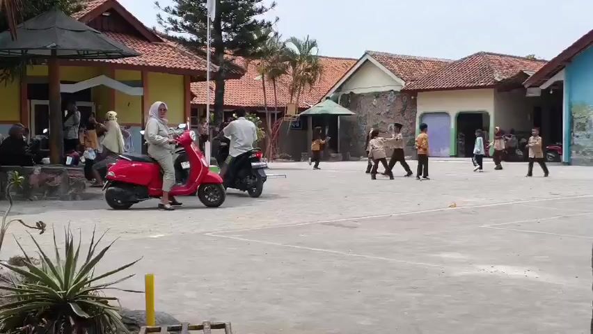 The video shows a courtyard with several buildings in the background. In the foreground, a red Vespa scooter is parked. A woman wearing a hijab and a light-colored dress is sitting on the scooter. To her left, a man in a white shirt and dark pants is standing next to a parked motorcycle. Further in the background, several children in uniforms are playing in the courtyard. Some children are walking, while others appear to be running or playing a game. The buildings have red tiled roofs and light-colored walls. The ground is paved with concrete. The sky is overcast.