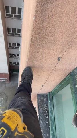 From a high angle, the video shows a person standing on a narrow ledge of a high-rise building. The person is wearing dark pants and black boots and is holding a yellow power drill. They are standing next to a window with a green frame. The building's exterior is textured and a peach-pink color. A wire runs vertically along the building's facade, secured by metal brackets. In the background, another part of the building can be seen, with multiple windows. The video appears to be taken from a first-person perspective, looking down and out from the building.