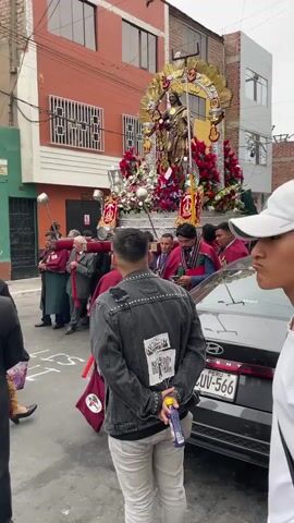 A religious procession is taking place on a narrow street lined with buildings. At the center of the frame, a statue of Jesus Christ is being carried on a large, ornate platform decorated with flowers. Several men, dressed in dark suits and burgundy robes, are carrying the platform. In front of them, other individuals are also wearing similar robes and appear to be part of the procession. On the right side of the frame, a silver car is parked, and two young men are standing near it, observing the procession. One of the young men is wearing a denim jacket with patches and light-colored pants, while the other is wearing a white baseball cap and a white t-shirt. A woman in a purple dress and a headscarf is visible on the far left, also watching the event. The buildings in the background have orange and gray facades, with some windows visible. The street appears to be paved, and there are some white markings on the ground. The overall atmosphere seems to be solemn and ceremonial.