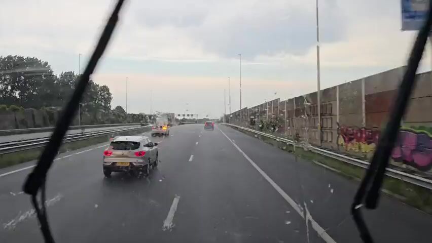 This video captures a view from the front of a vehicle driving on a highway in Zoetermeer, Netherlands. The weather appears to be overcast and possibly raining, as evidenced by the water droplets on the windshield and the wet road surface. The sky is a mix of gray clouds and lighter patches, suggesting it's either early morning or late afternoon.

The highway has multiple lanes, with several cars visible in front of the vehicle. One car in the foreground, a white SUV, has its brake lights illuminated. Further down the road, other vehicles can be seen, including a red car and a lighter-colored car. A large sound barrier, constructed from concrete panels, lines the right side of the highway. Some of these panels are marked with graffiti. On the left side of the highway, trees and greenery are visible, with some appearing to have been recently trimmed. Streetlights are spaced along the sides of the road, and some appear to be illuminated, further hinting at the time of day.

The activity taking place is vehicular traffic on a highway. The overall mood conveyed is one of travel, with the wet weather adding a slightly somber or subdued tone to the scene. There is no visible text in the video.