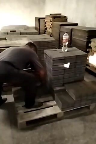 A man in dark clothing is in a warehouse, stacking dark, rectangular objects onto wooden pallets. The objects appear to be bricks or paving stones. In the background, there are stacks of similar objects. On top of one of the stacks of objects, there is a plastic bottle of water. The man is in the process of moving and stacking these objects. The lighting is dim, suggesting artificial light.