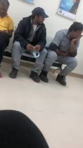 Three adult men are seated on a metal and black plastic bench in an indoor waiting area. To the left, a man in a yellow polo shirt sits with his hands in his lap, gazing downward. In the center, a man wearing a dark blue baseball cap and a black jacket is holding a blue and white face mask in his hands, with his attention directed towards the man to his right. The man on the right, dressed in a gray jumpsuit, has his elbow resting on his knee and his hand supporting his head, creating the impression of fatigue or contemplation. All three men are wearing dark shoes.

The waiting area has a clean, light-colored tiled floor and white walls. Behind the seated men, there are posters visible on the wall. The recording appears to be taken from a lower angle, showing part of a black object in the foreground, possibly a piece of clothing.