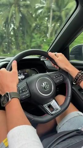 The video begins with a first-person perspective of someone driving a car. The driver's hands are visible on the steering wheel, which has an MG logo in the center. They are wearing a watch on their left wrist. The car's dashboard display is partially visible. The exterior view through the windshield shows a road lined with numerous palm trees. The sky is overcast. Another white car is visible ahead on the road. The car continues to drive along the road, which curves to the right. The perspective shifts to show the view through the rearview mirror, reflecting the driver's face and the road behind. The camera then shows the road ahead again, with more palm trees and a winding road. Another white car is visible driving in the opposite direction. The video ends with the car continuing to drive down the palm-lined road.