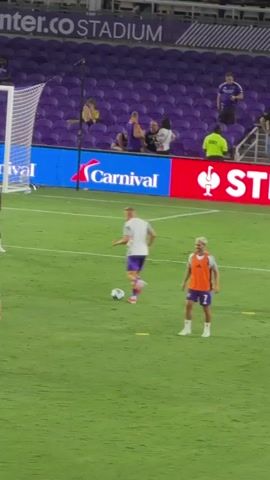 The video shows a soccer field with players in orange vests and purple shorts practicing. Some players are running and kicking a soccer ball, while others are standing and observing. There are spectators in the background, and a large advertising banner is visible behind the field. The environment appears to be a stadium.