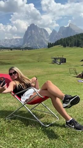 A blonde woman wearing sunglasses, a black tank top, white ripped shorts, and black sneakers is sitting in a red lawn chair and taking a selfie with her phone. The woman is in the foreground and is taking the selfie with her right hand. She is looking at the camera on her phone. The woman's legs are crossed. Behind the woman is a grassy field with mountains and a ski lift in the background. The sky is blue with some white clouds.