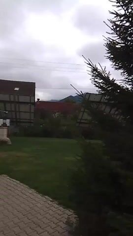 A handheld video camera pans from a paved walkway and green grass towards a view of a residential area with houses and vegetation under a cloudy sky. In the foreground, a tree branch is visible on the right. In the middle ground, a portion of a house with a brown roof and a red tiled roof can be seen, along with trees and bushes. In the background, mountains are visible, shrouded in mist or clouds. Electrical wires are strung across the sky. The scene appears to be set in a rural or suburban environment.