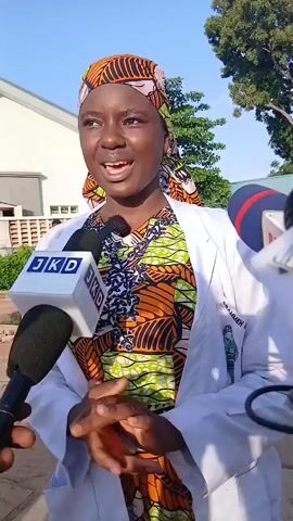 A young woman wearing a white lab coat over a colorful patterned dress and a headscarf is being interviewed by two microphones. One microphone is closer to her, with the logo JKD visible. The other microphone is to her right. She appears to be outside, with a building and trees in the background. Her hands are clasped in front of her. The sun is shining.