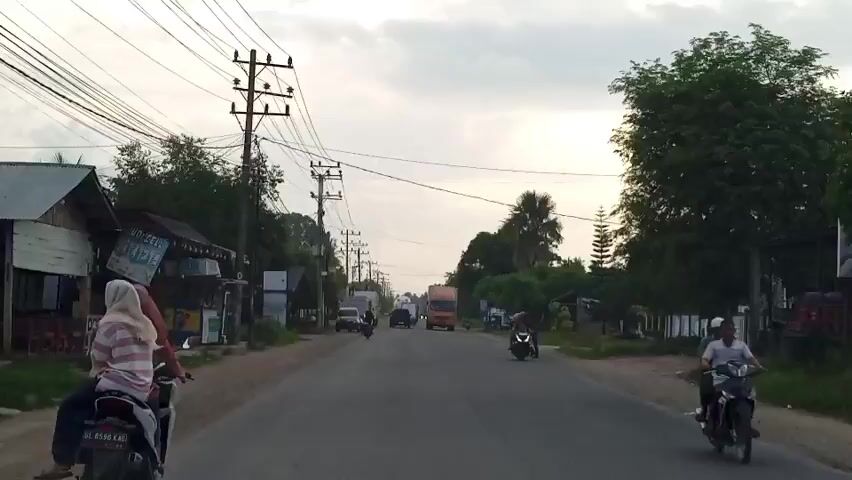 The video shows a two-lane road with a person on a motorbike in the foreground. There are buildings and utility poles lining the road, as well as trees and vegetation. Several vehicles, including trucks, cars, and motorbikes, are visible on the road. The sky is overcast. A person is crossing the road in front of an approaching truck. The overall impression is that of a rural or semi-urban road scene.