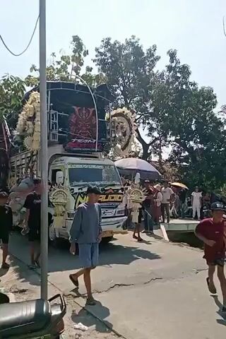 A white truck with extensive decorations, including large white floral arrangements on either side of the front, a large samurai mask graphic on top, and "TEAM AUDIO" written on it. The truck is also adorned with various smaller decorative elements and lights. A group of people, mostly young males wearing casual clothing, are walking alongside and in front of the truck. Some appear to be participating in a parade or procession. The environment appears to be an outdoor street with trees and buildings in the background, under a bright sky.