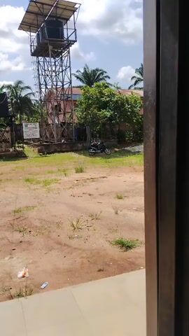 The video shows a view from a window, looking out onto a sandy area. In the distance, there is a metal water tower with a black tank on top, next to another smaller black tank. There is a building, some trees, and a motorcycle parked in the background. The sky is blue with some clouds. There is some trash on the ground in the foreground.