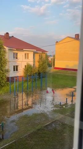 Two children, one in a red shirt and the other in a red shirt with white shorts, are playing in a large puddle in an outdoor recreational area. The puddle has formed on a grassy area, partially covering a set of blue playground equipment. Behind the playground, two residential buildings are visible, one with a white facade and a red roof, and another with a yellow facade. The sky is blue with scattered white clouds, and the sun appears to be setting, casting a warm glow on the buildings.