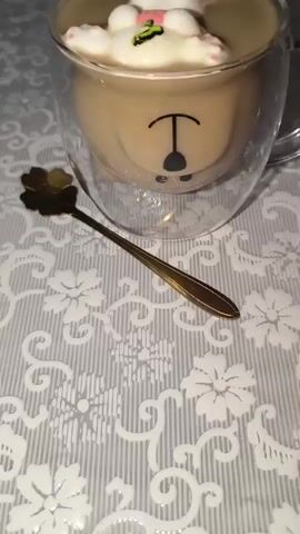 This is a close-up video of a hot beverage in a clear glass mug, decorated with a marshmallow bunny. The mug has a double-wall design and appears to have a cartoon bear face printed on the inside. The bunny, made from marshmallows, is floating on top of the drink. It has pink accents on its ears, eyes, and paws, and is holding a small flower with a green stem and pink petals. A golden spoon with a floral-shaped head rests on the table next to the mug. The background is a textured tablecloth with a white floral pattern. The lighting suggests it could be daytime, and the overall mood is whimsical and charming, likely intended for a festive occasion like Easter. No text is visible in the media.