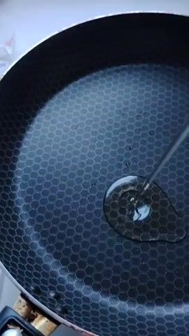 A close-up, high-angle shot shows cooking oil in a hexagonal-patterned frying pan. The pan is on a stovetop, and a glass lid is next to it. The oil is shimmering, indicating that the pan is heating up. There are no people visible in the frame.
