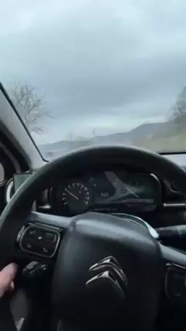 A video shows the view from inside a moving car, focusing on the driver's hands on the steering wheel and the dashboard. The car is a Citroën, recognizable by its logo on the steering wheel. The dashboard displays a speed of around 60 kilometers per hour, which fluctuates slightly during the short video. The car is traveling on a road, with a landscape visible through the windshield, featuring hills and a cloudy, overcast sky. A mobile phone is mounted to the left of the steering wheel. The interior of the car appears clean and modern. The driver's hands occasionally adjust their grip on the steering wheel, suggesting they are navigating the vehicle.