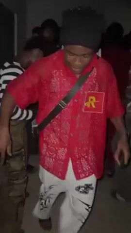 The video shows a group of people dancing in what appears to be an indoor venue.  The primary focus is on a person in the center wearing a red, slightly translucent shirt with a square emblem on the left side, white patterned pants, a black beanie, and a black crossbody bag. This person is actively dancing, moving fluidly with the music. Other people are visible in the background, also dancing or moving to the music. Some are wearing distinctive clothing, like a black and white striped shirt. The setting seems dimly lit, but it is clear that the people are enjoying themselves in a festive atmosphere.
