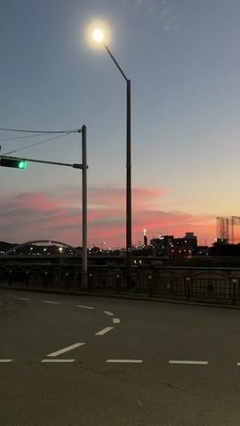 The video shows a street scene at dusk. A street light is illuminated, casting a bright glow. A traffic signal with a green light is visible, along with two traffic signs. One sign shows a speed limit of 60, and the other has an arrow indicating a turn. The sky displays a beautiful gradient of colors, with pink and orange hues from the sunset. In the distance, a bridge spans across a body of water, and buildings can be seen on the horizon. The road in the foreground has white dashed lines marking the lanes, and there appears to be a slight curve to the road. A faint silhouette of what looks like a cross is visible on a building in the background. The overall atmosphere is peaceful and serene, capturing the transition from day to night.